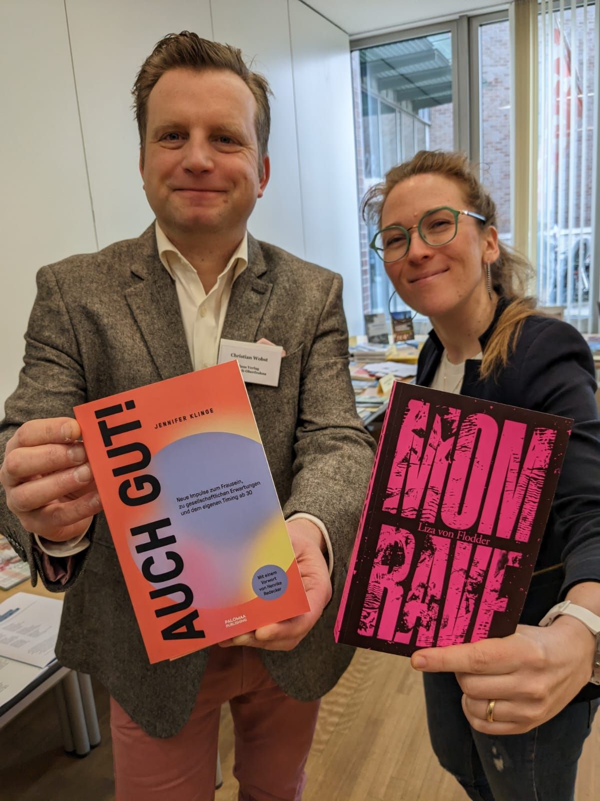 Ein Mann und eine Frau stehen in einem Büro und halten jeweils ein Buch in die Kamera. 