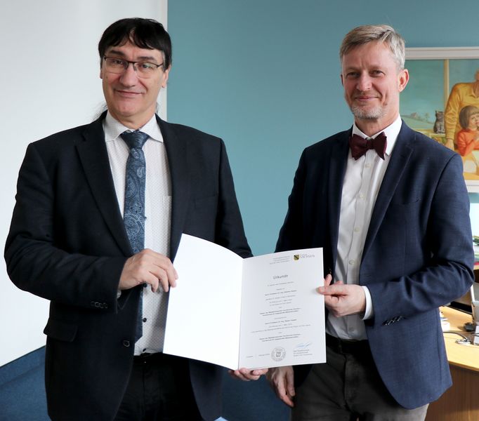 Rektor und Staatssekretär halten Urkunde und blicken in die Kamera.