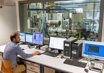 Ein Wissenschaftler sitz vor Monitoren die verschiedene Messdaten anzeigen. Im Hintergrund, hinter einer Glasscheibe, ist ein Motorenprüfstand zu sehen.