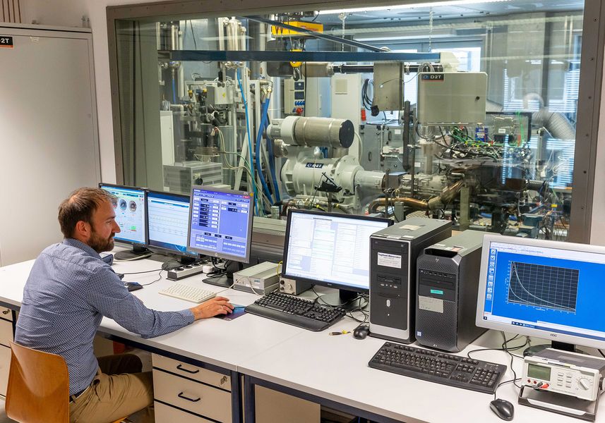 Ein Wissenschaftler sitz vor Monitoren die verschiedene Messdaten anzeigen. Im Hintergrund, hinter einer Glasscheibe, ist ein Motorenprüfstand zu sehen. 