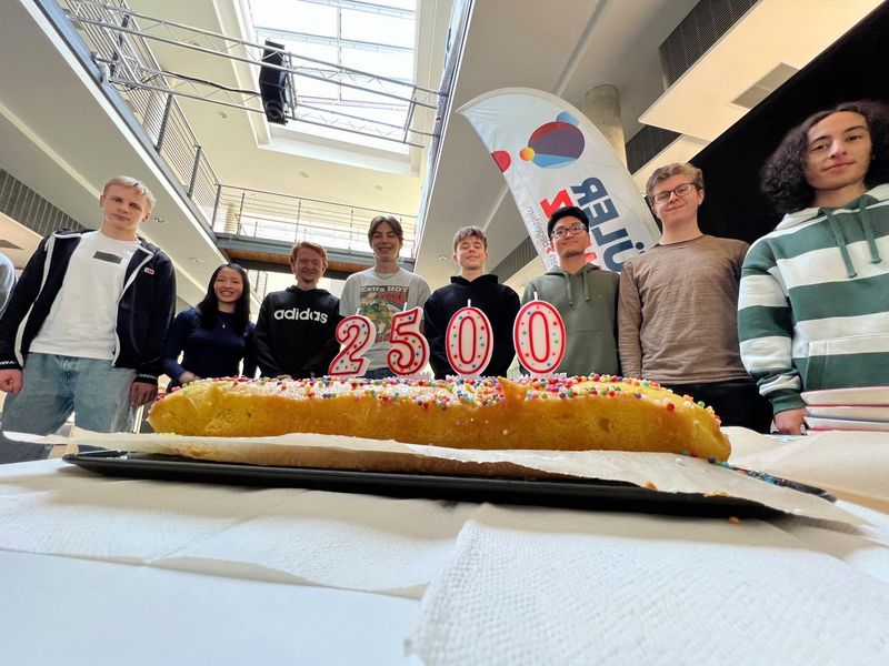 Im Vordergrund ein Kuchen mit einer 2500, im Hintergrund Schüler. 
