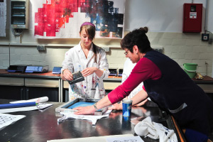 Foto: Zwei Studierende bearbeiten an einem Arbeitstisch einen Stoff mit Farbe unter Anleitung einer Lehrenden.