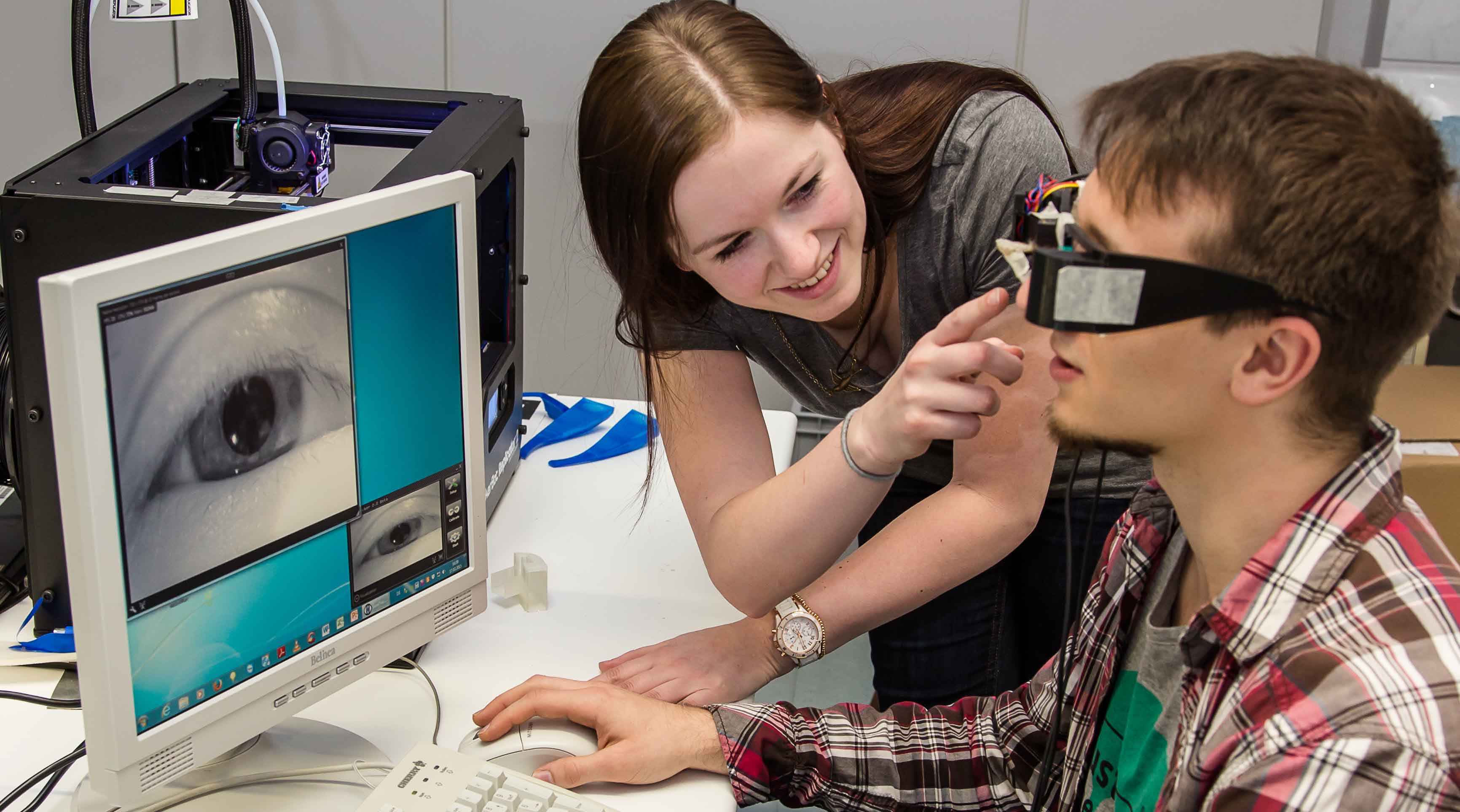 Photo: A student wears glasses with a camera. His eye can be seen on a screen in front of him. A female student stands next to him and assists. (source: whz/H. Gerischer)