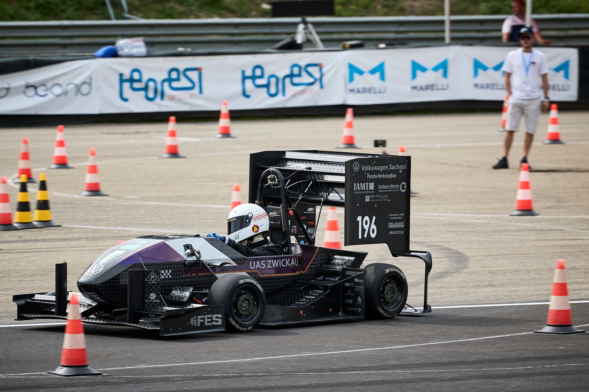 Im Zentrum des Bildes ist der Rennwagen des WHZ Racing Teams zu sehen. Er fährt auf einer Rennstrecke. Im Hintergrund sieht man orangene Hütchen und eine Person.