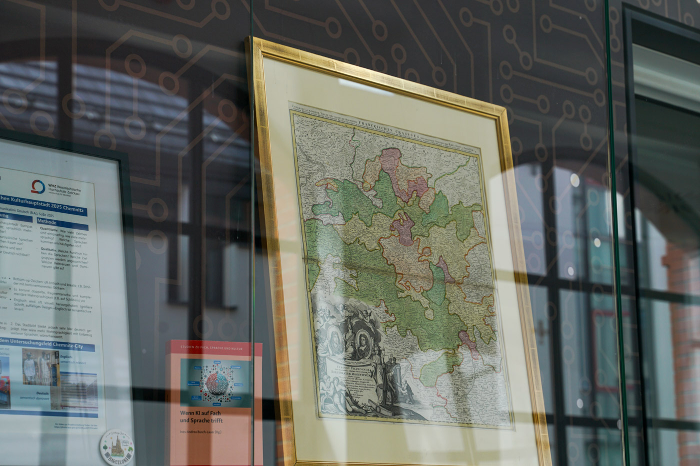 Auf dem Bild sieht man die ausgestellte Frankenkarte seitlich in der Vitrine stehen. Sie befindet sich in einem goldenen Rahmen. Links davon stehen ein Fachbuch zu KI und Sprache und ein Bilderrahmen.