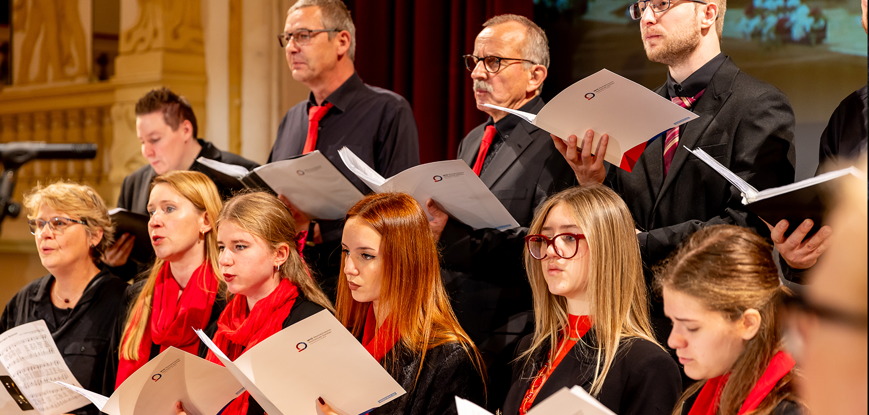 Chormitglieder stehen mit Notenheft und festlicher Klediung auf einer Bühne. 