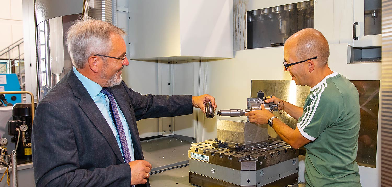 Foto: Titelbild Forschungsprofile. Zwei Professoren stehen an einem technischen Aufbau. Der linksstehende Professor hält ein Werkstück in der linken Hand. Der rechtsstehende Professor richtet ein Gerät auf das Werkstück aus.