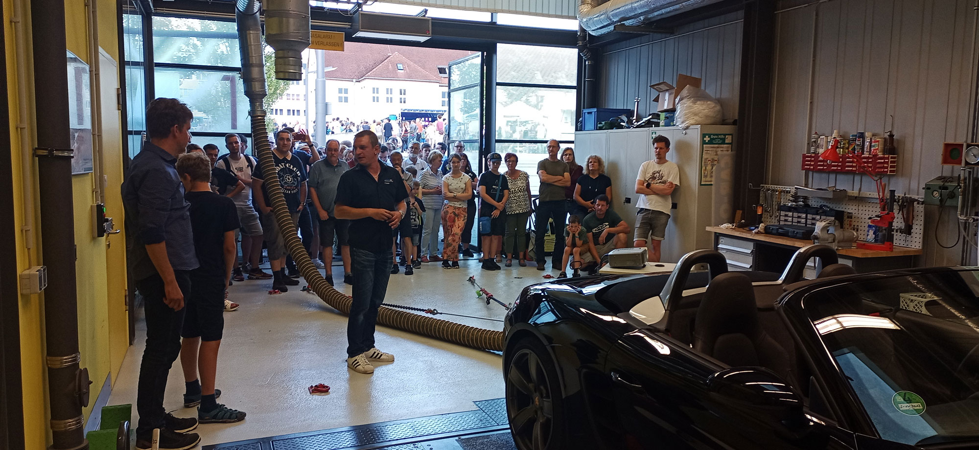 Foto: zu sehen ist ein Auto auf einem Rollenprüfstand mit angeschlossenem Abgasschlauch und viele interessierte Gäste, die gespannt zuschauen | Quelle:WHZ