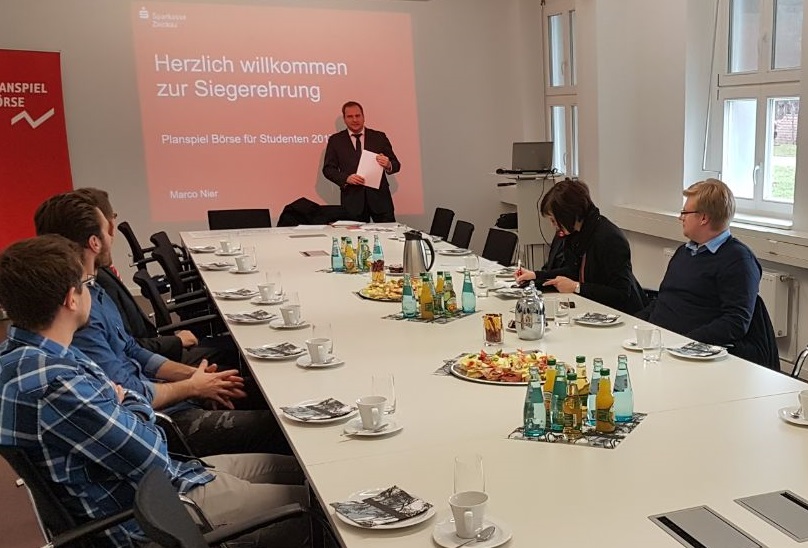 Foto: In einem Besprechungsraum sitzen Teilnehmer des Planspiel Börse. Ein Referent präsentiert eine Präsentation mit der Aufschrift "Herzlich willkommen zur Siegerehrung".