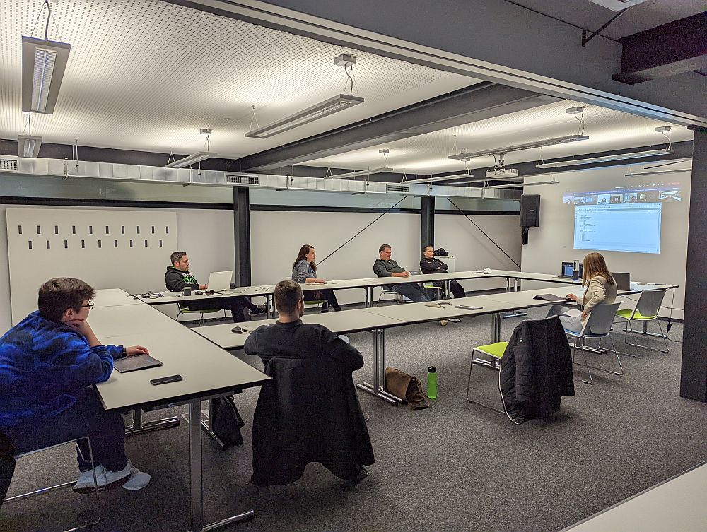 Foto: In einem großen Beratungsraum stzen einige Menschen weit auseinander. Auf einer Leinwand sind weitere Personen zugschalten. 