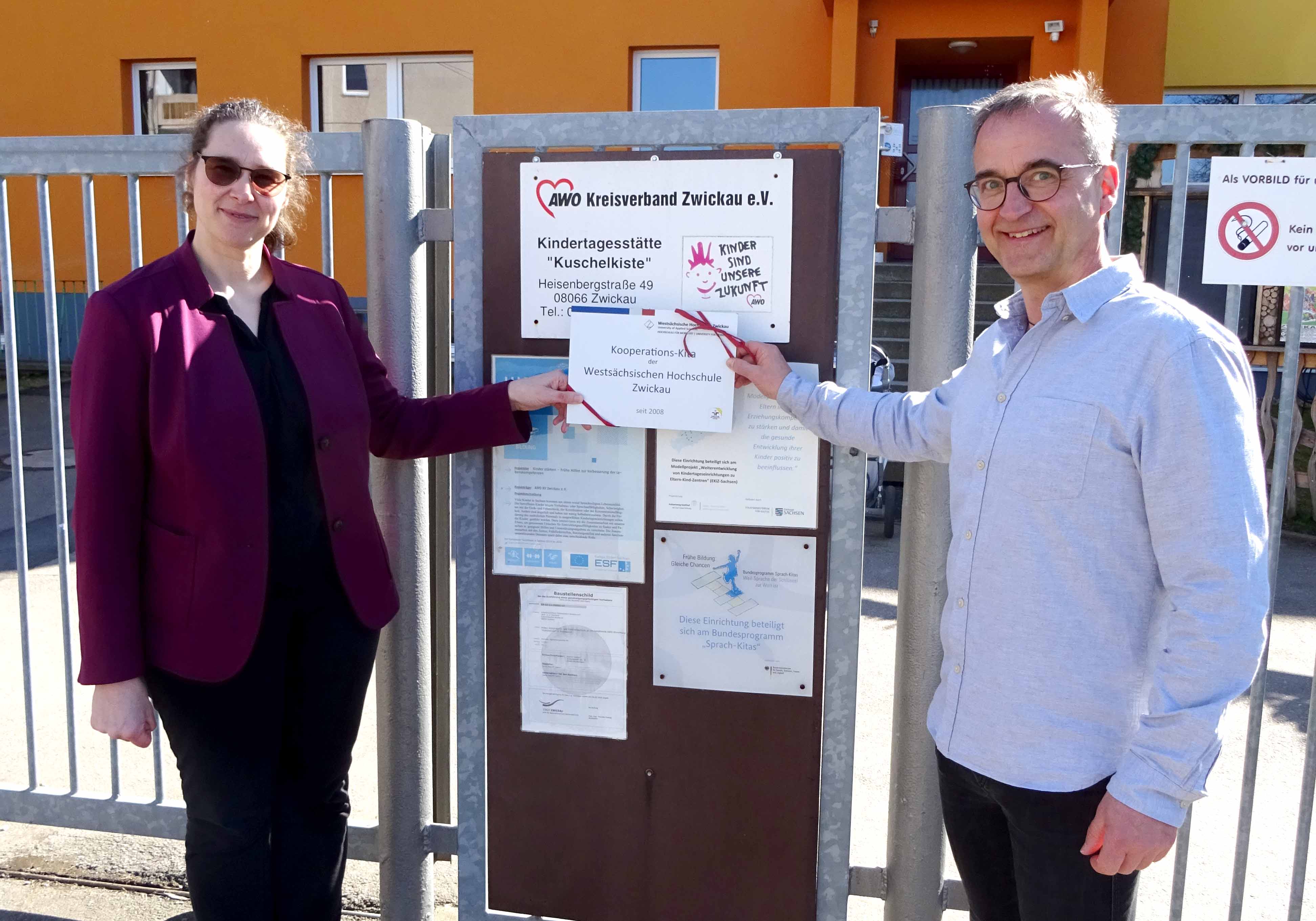 Prof. Anke Häber, Beauftragte für die Familiengerechte Hochschule und Einrichtungsleiter Jens Kluge vor dem Eingang der Kita. In der Hand halten Sie ein Schild mit der Aufschrift Kooperationskita der Westsächsischen Hochschule Zwickau. 