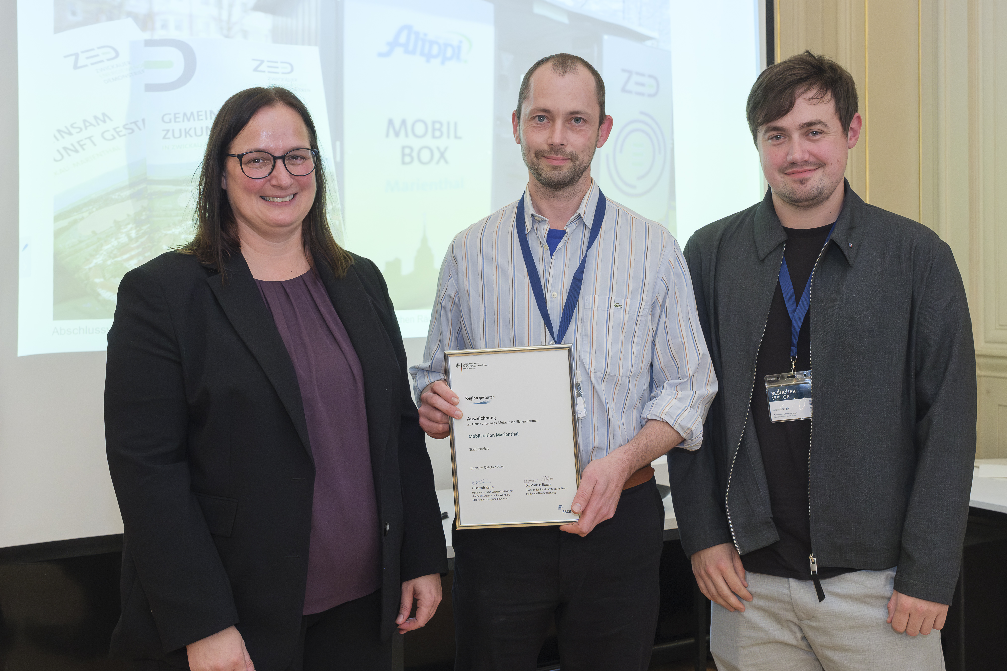 Drei Personen lächeln in die Kamera, die mittlere Person hält eine Urkunde hoch. 