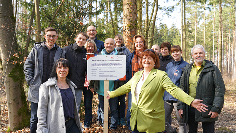 Personen stehen im Wald und blicken fröhlich in die Kamera, zwischen Ihnen ein Schild mit einem Hinweis auf die Pflanzaktion.. 