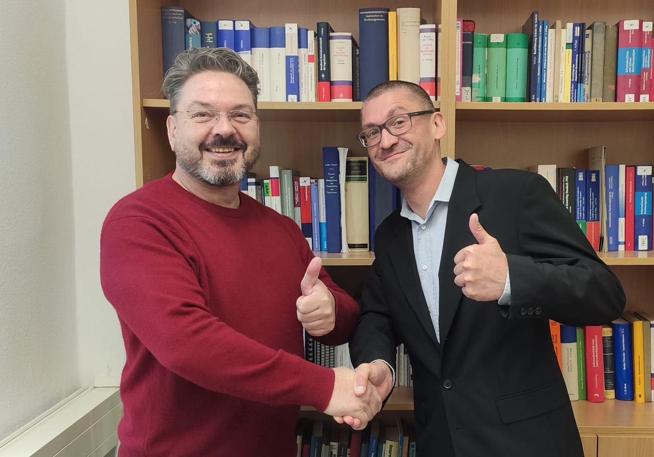 Zwei Personen blicken freudig in die Kamera, im Hintergrund ein Regal mit Bürchern. 