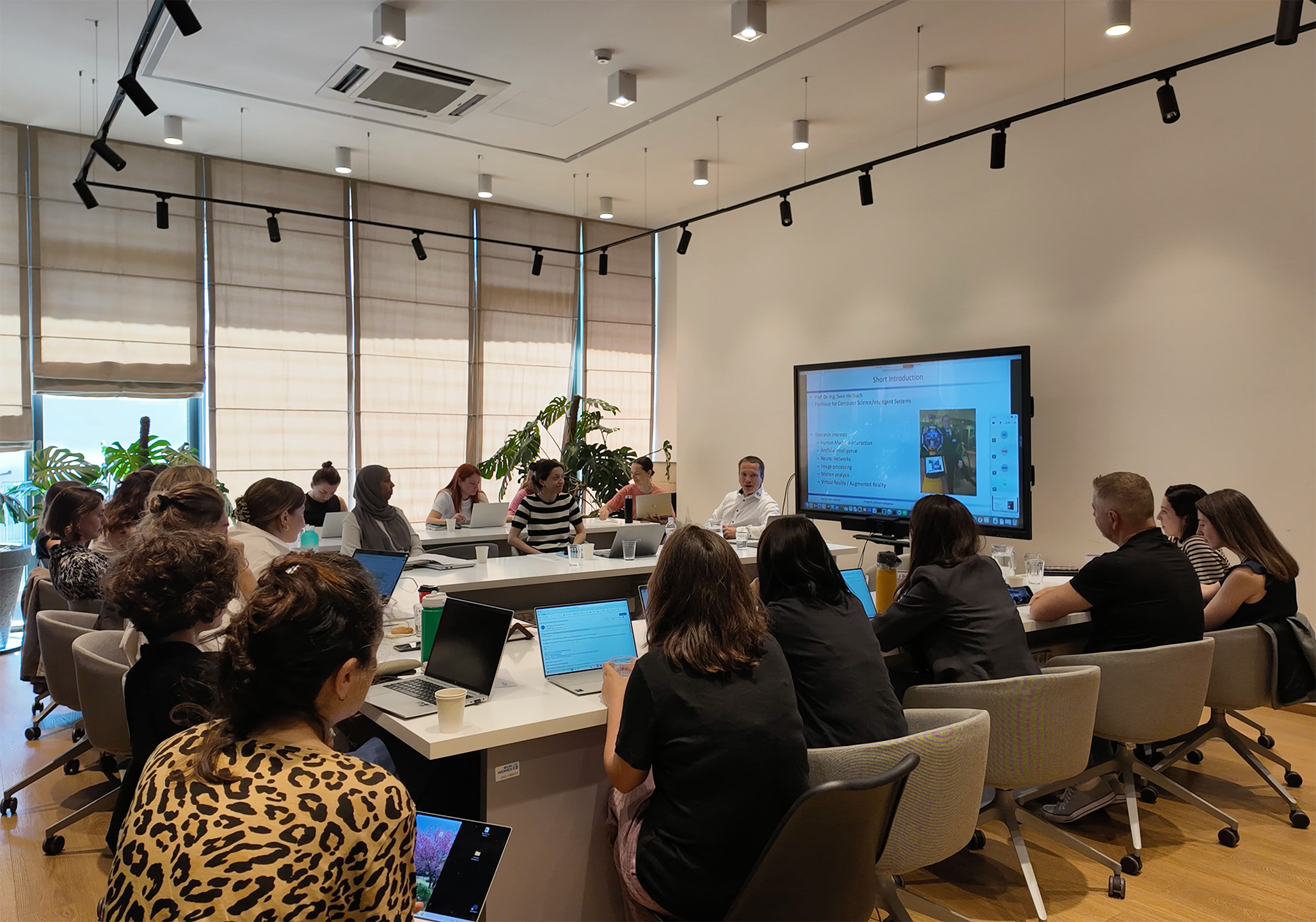 Teilnehmende sitzen um einen großen Tisch und hören zu, im Hintergrund läuft eine Präsentation auf einem Bildschirm. 