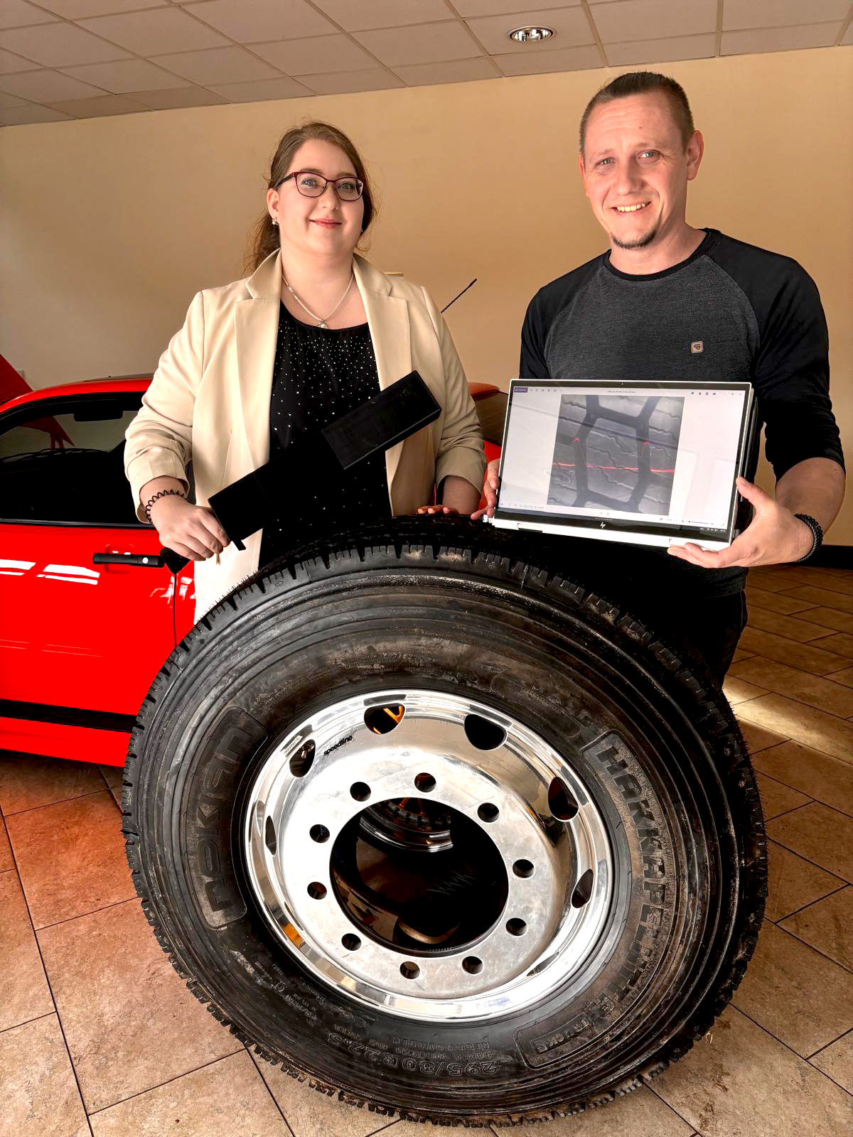 Zwei Personen stehen in einer Garage hinter einem LKW-Reifen und präsentieren ein Messsystem. . 