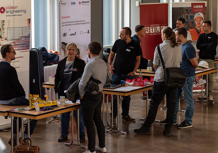 kleine Messestände von Unternehmen in einer Aula an denen sich Menschen unterhalten