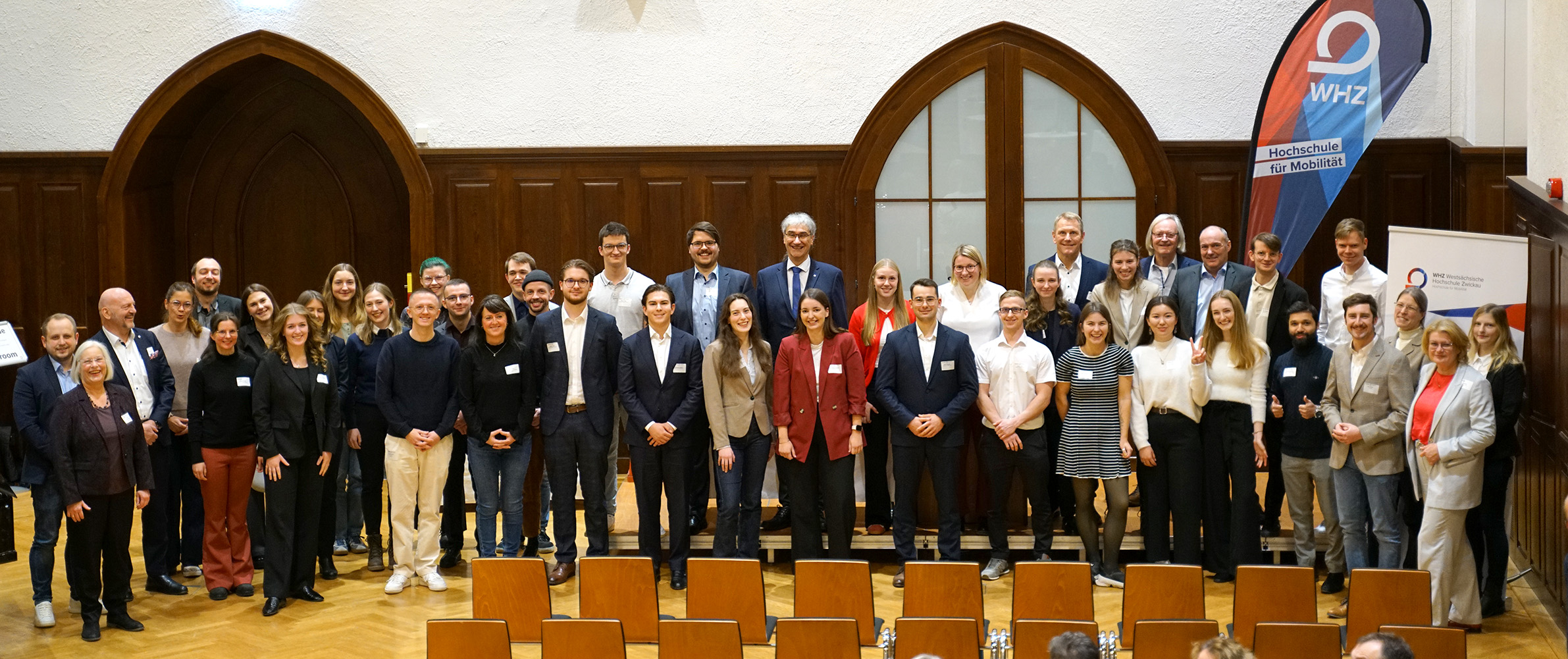 Eine große Gruppe von Personen in festlicher Kleidung steht in einem Saal. 