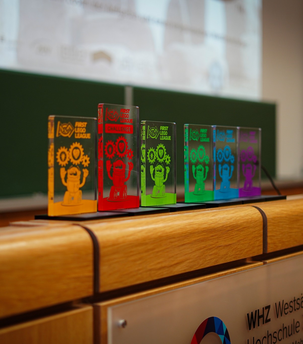 Sechs Pokale stehen aufgereit und in unterschiedlichen Farben vor einer Tafel im Hörsaal. Sie sind mit einem Roboter bedruckt und flach.