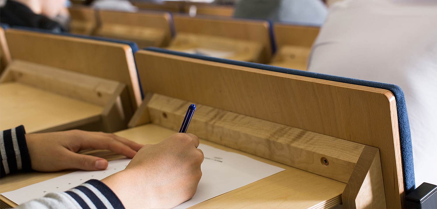 Eine Hand hält einen Stift. Vor der Hand ein beschriebenes Papier auf einem Hörsaalpult. 