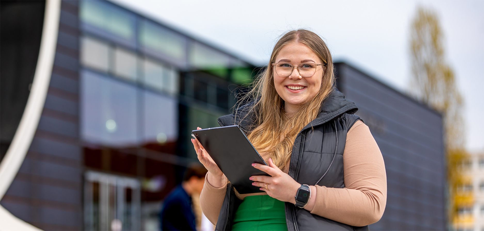 Eine Studenierende steht mit einem Tablett vor dem August-Horch-Bau und blickt in die Kamera 