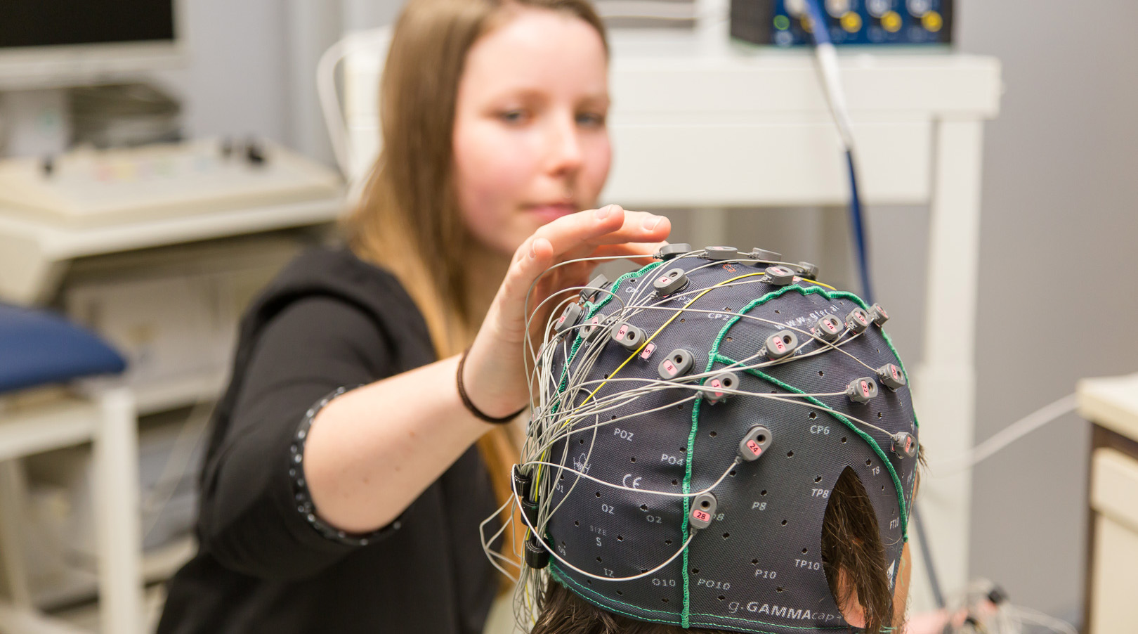 Foto: Zwei Studierende in einem Forschungslabor. Diese führen Untersuchungen mit einer Elektroenzephalografie durch. Einer der Studierenden stellt den Probanden dar.
