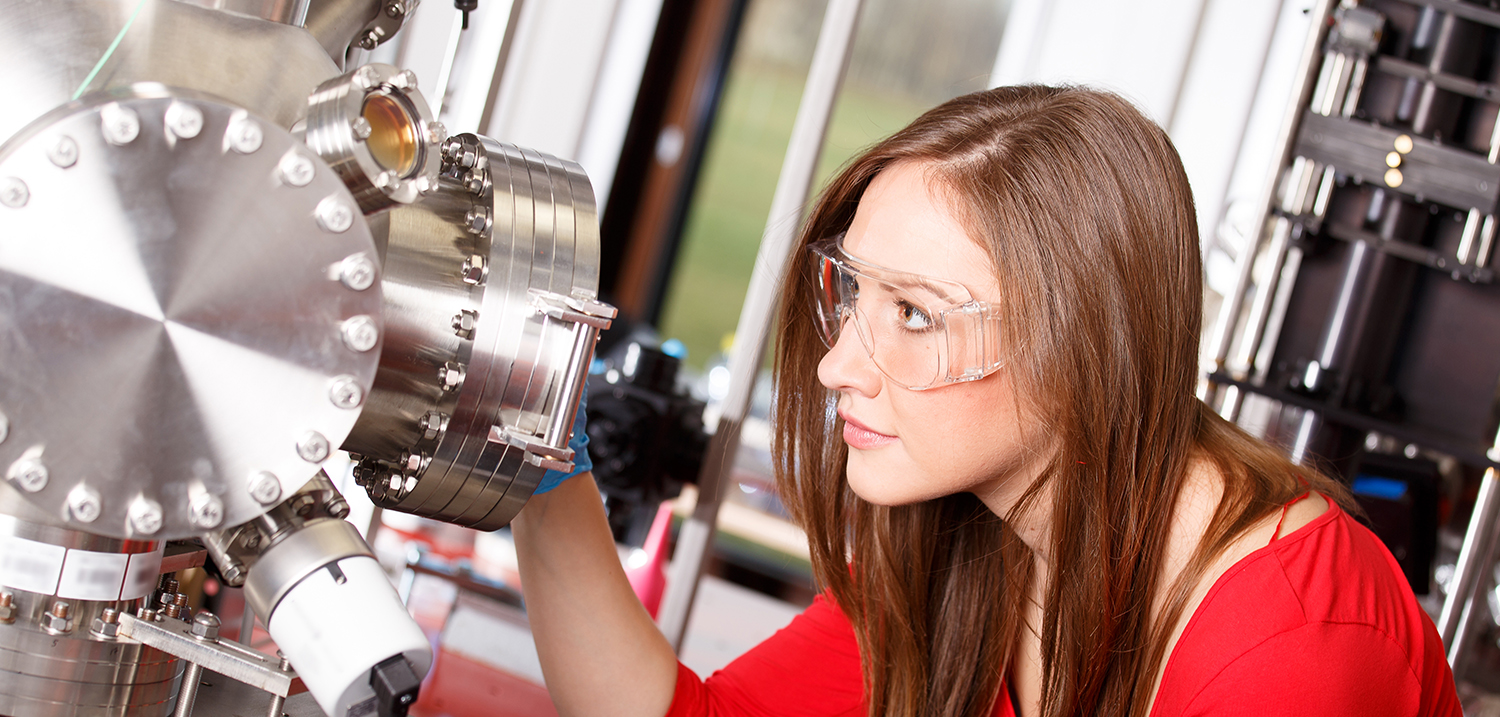 Eine Frau mit Schutzbrille betrachtet ein Mikroskop. 