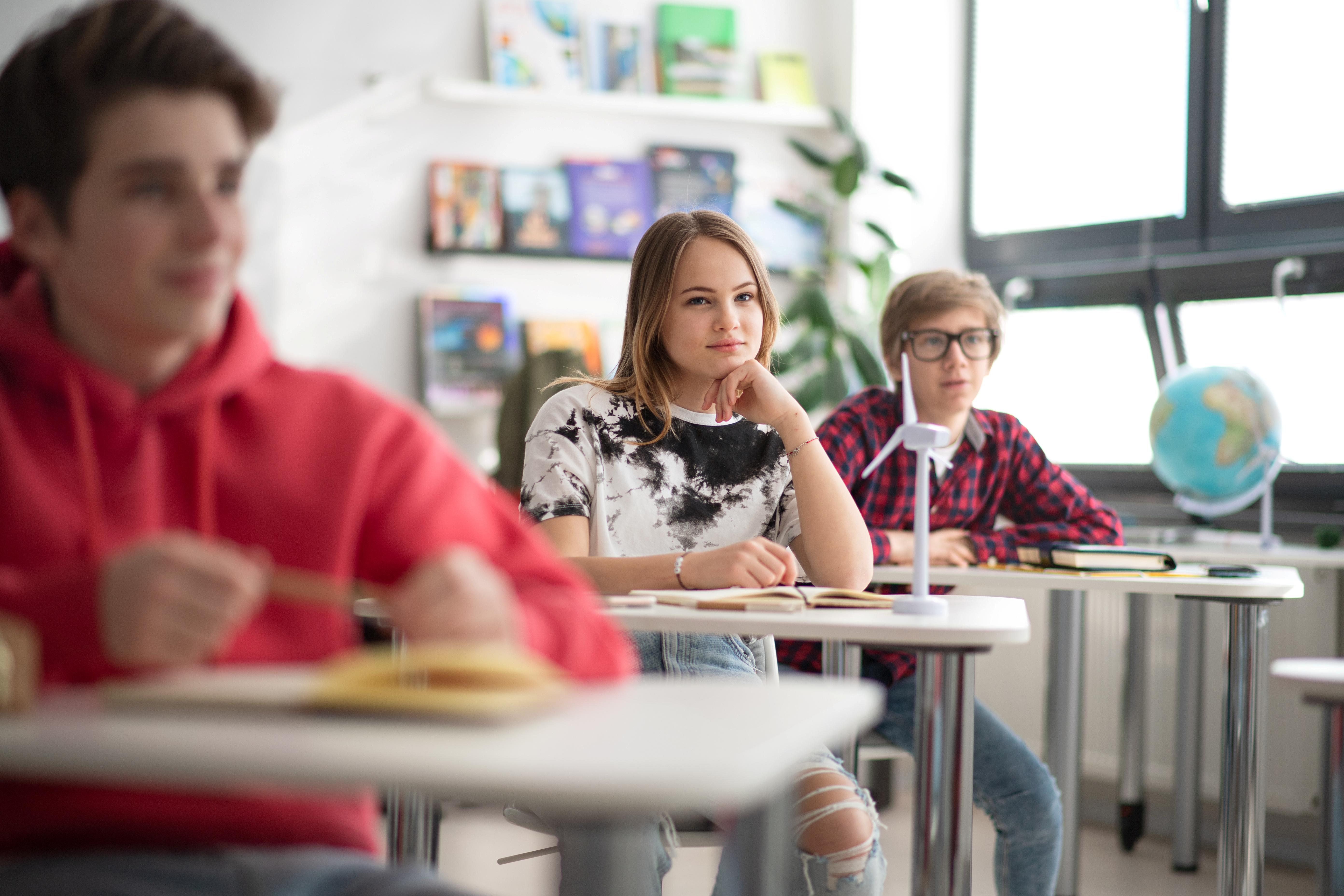 Schülerinnen und Schüler sitzen in einem Klassenraum und schauen interessiert in Richtung der Tafel. Im Fokus steht ein Mädchen, das ihren Kopf auf ihrer Hand abstützt.